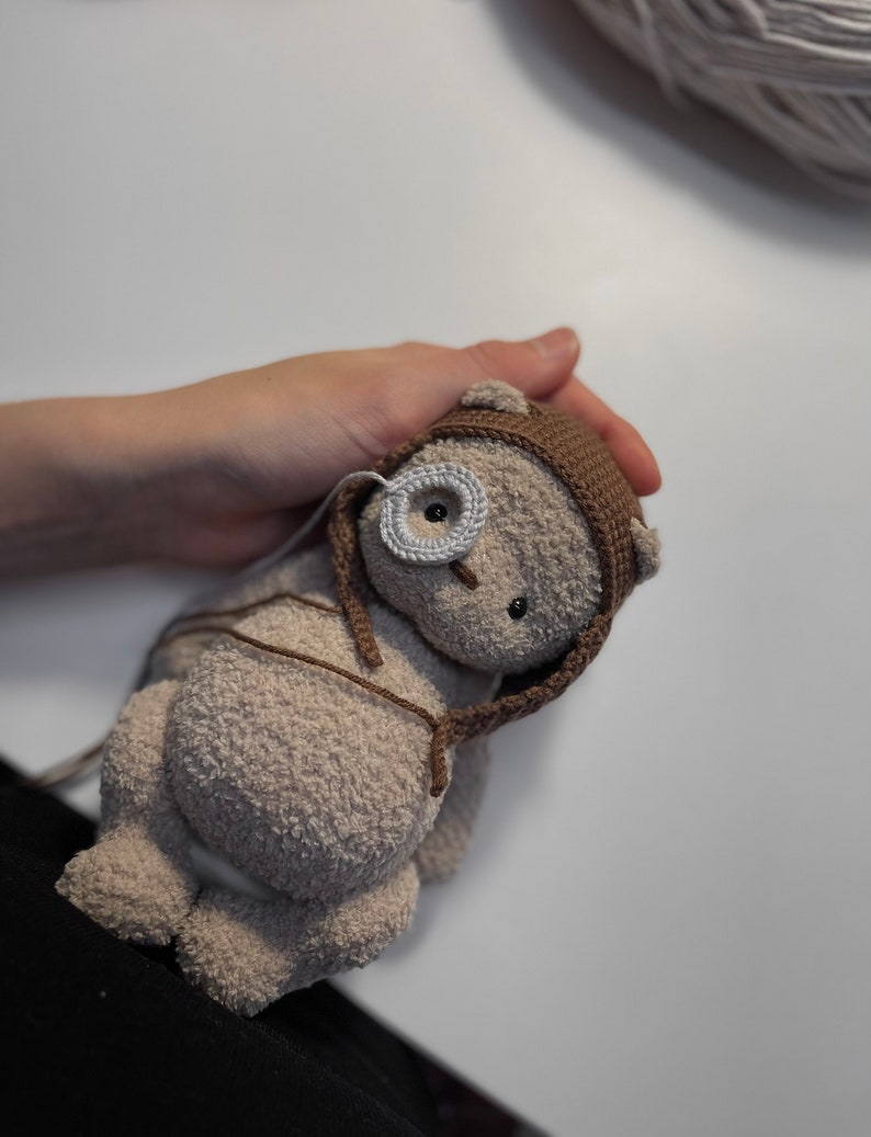 May include: A crocheted brown teddy bear with a brown and white crocheted hat. The bear has a white button eye and a black button nose.