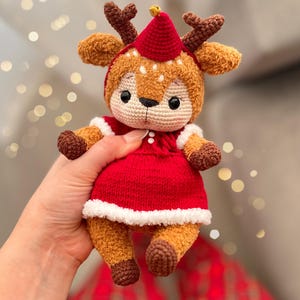 May include: A handmade crochet deer doll wearing a red dress with white trim and a matching red hat. The deer has brown antlers, ears, and hooves, with a beige face and a small gold bell on its hat. The doll is approximately 7 inches tall.