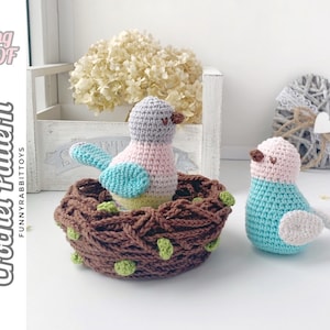 May include: Two crocheted birds, one in a brown nest. The bird in the nest is pink, gray, and blue. The other bird is blue and white. The birds are sitting on a white surface.