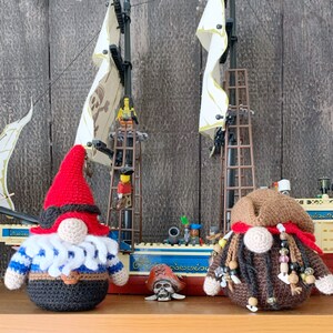 May include: Two crocheted gnomes, one in red and white and the other in brown and white, are standing in front of a toy pirate ship. The ship has white sails and a brown wooden hull. The gnomes are wearing pirate hats and have beards.