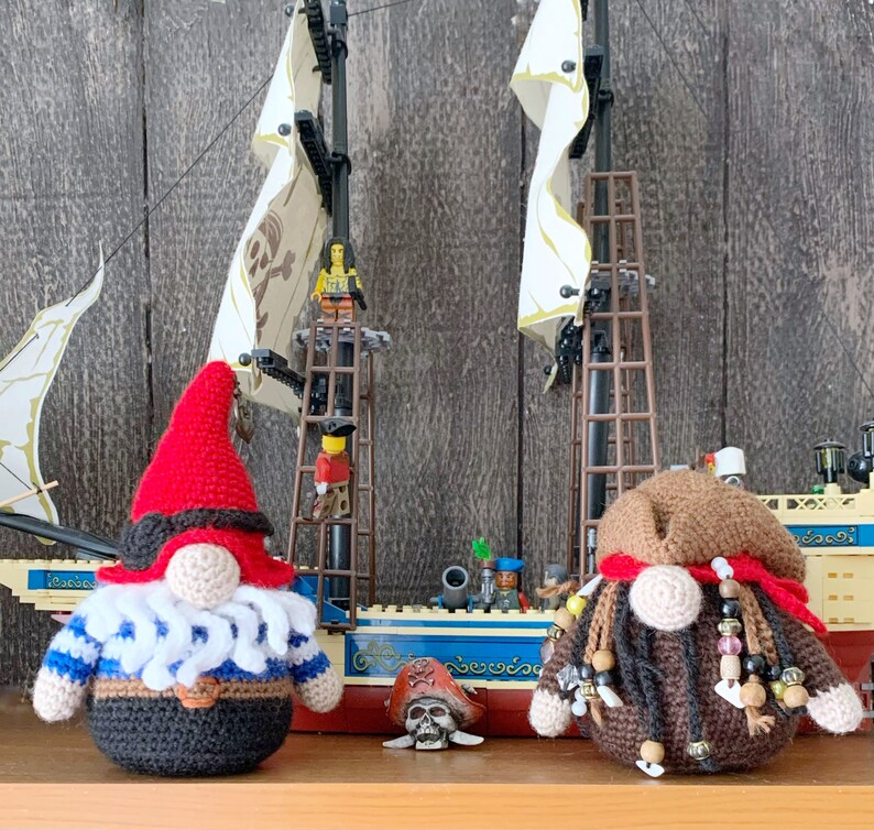 May include: Two crocheted gnomes, one in red and white and the other in brown and white, are standing in front of a toy pirate ship. The ship has white sails and a brown wooden hull. The gnomes are wearing pirate hats and have beards.