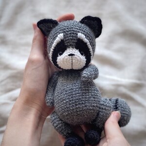 May include: A crocheted gray raccoon stuffed animal with black eyes and a white nose and mouth. The raccoon is being held by two hands.