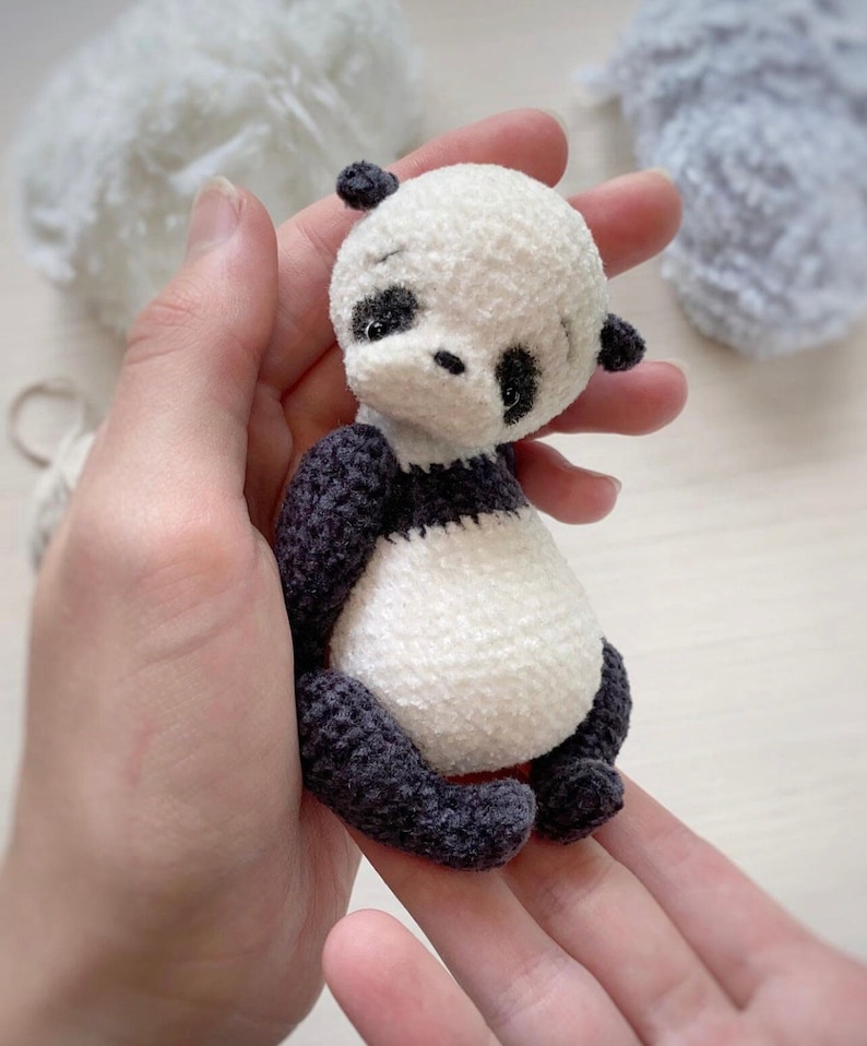 Peut inclure: Un petit ours en peluche panda crochet&eacute; avec de la fourrure noire et blanche. Le panda est assis dans une main, la t&ecirc;te inclin&eacute;e sur le c&ocirc;t&eacute;.