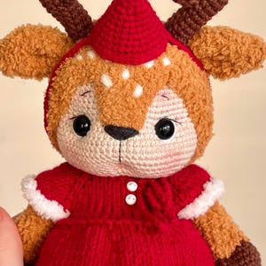 May include: A handmade crochet reindeer doll with a red dress and hat. The doll has brown antlers, a beige face with black eyes, and white spots. A small gold bell adorns the top of the hat. The doll is a festive holiday decoration.