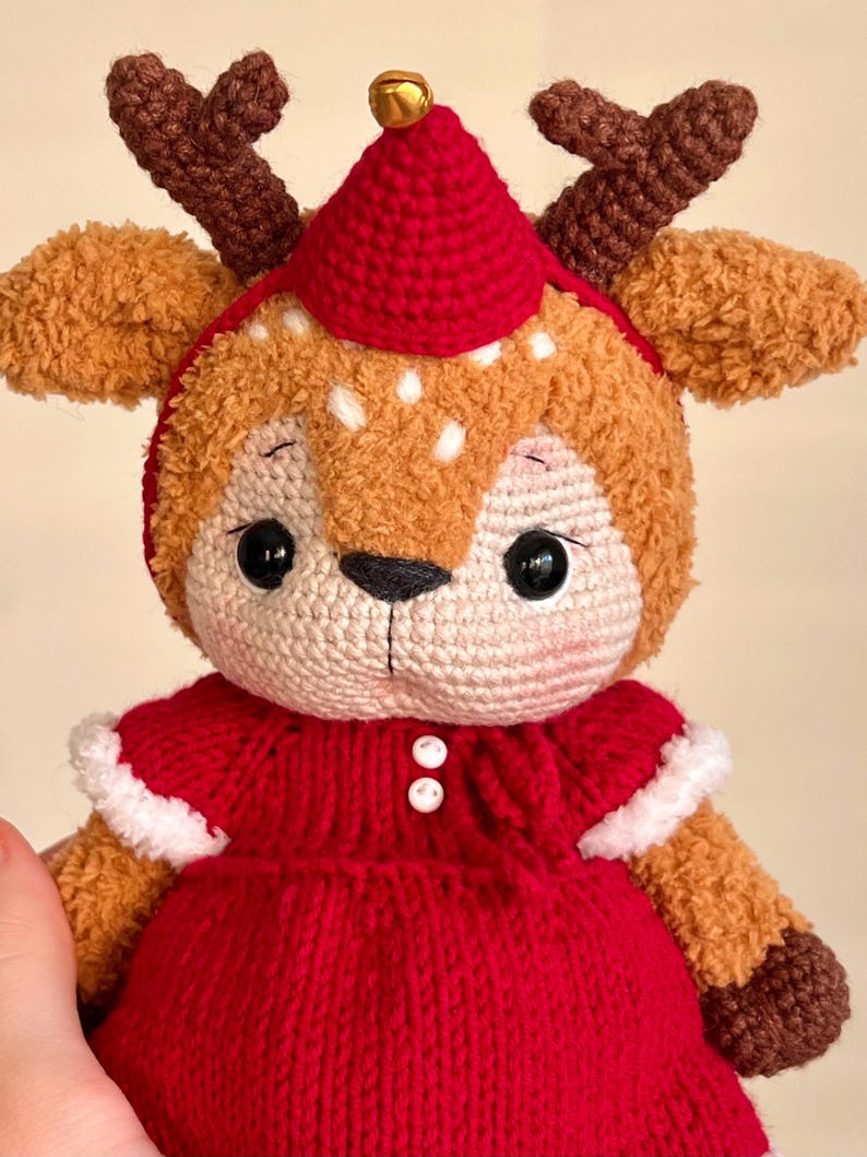 May include: A handmade crochet reindeer doll with a red dress and hat. The doll has brown antlers, a beige face with black eyes, and white spots. A small gold bell adorns the top of the hat. The doll is a festive holiday decoration.