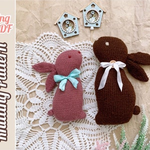 May include: Two knitted bunny rabbit toys, one brown and one pink, with white and blue ribbon accents. The bunnies are sitting on a white crocheted doily.