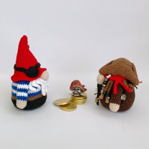 May include: Two crocheted gnomes, one with a red hat and blue and white striped shirt, the other with a brown hat and red scarf. Both gnomes have a white beard. A small pirate skull with a red hat is in front of the gnomes.