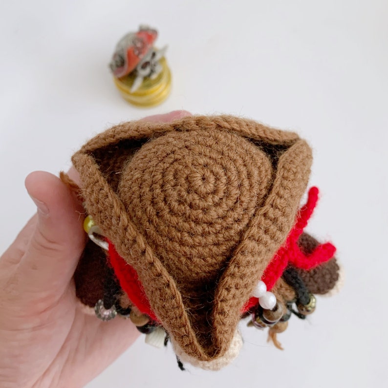 May include: A brown crocheted pirate hat with a red bandana and white beads. The hat is being held by a hand.