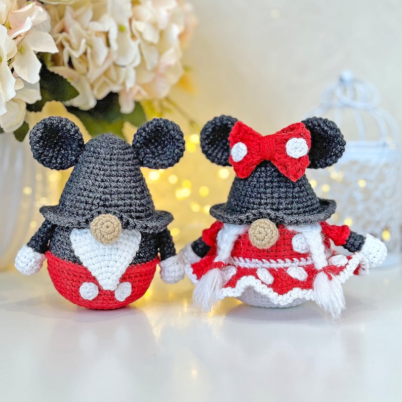 May include: Two crocheted gnome ornaments, one with a red and white polka dot dress and a red bow, and the other with a red and black Mickey Mouse inspired outfit.