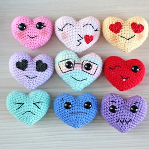 May include: Nine crocheted heart-shaped plush toys with different facial expressions. The hearts are in various colors, including pink, yellow, purple, blue, and red.