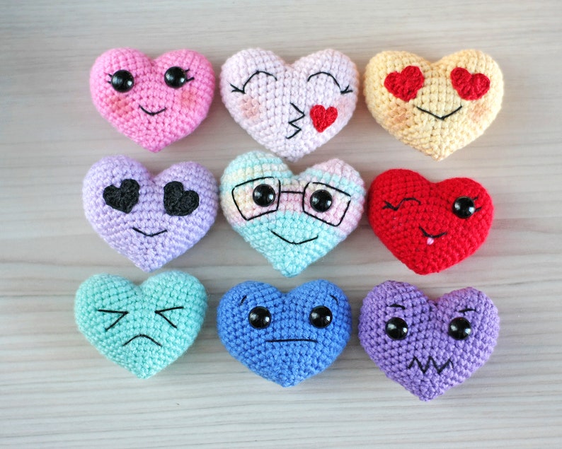 May include: Nine crocheted heart-shaped plush toys with different facial expressions. The hearts are in various colors, including pink, yellow, purple, blue, and red.
