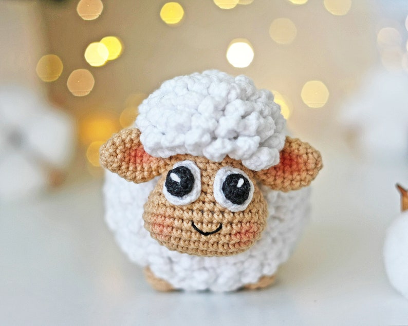 Pu&ograve; includere: Un giocattolo di pecora in crochet bianco con orecchie marroni e occhi neri. La pecora ha una bocca sorridente ed &egrave; seduta su una superficie bianca.