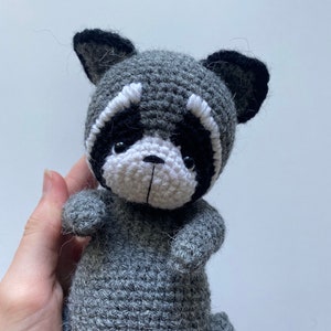 May include: A gray crocheted raccoon plush toy with black ears and a white face. The toy has black eyes and a small black nose.