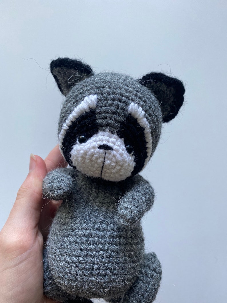 May include: A gray crocheted raccoon plush toy with black ears and a white face. The toy has black eyes and a small black nose.