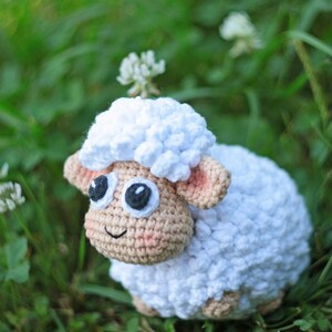 Pu&ograve; includere: Un giocattolo di pecora in crochet bianco con occhi neri e naso rosa, seduto in un ciuffo di erba verde.