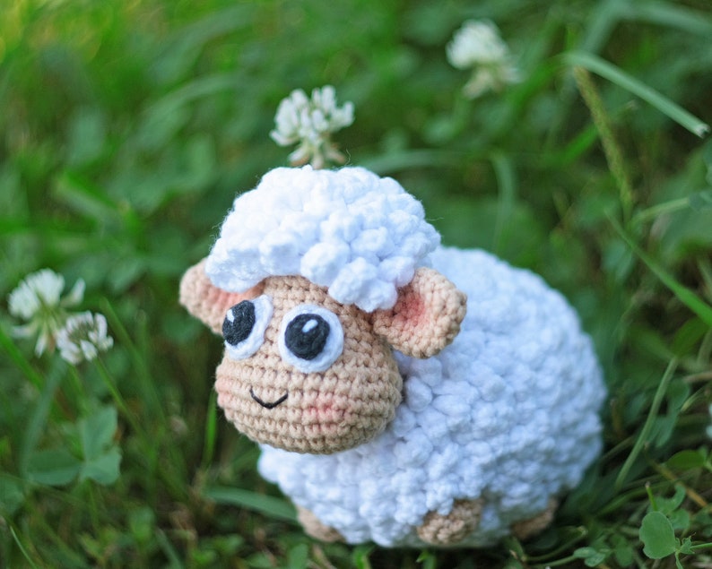 Pu&ograve; includere: Un giocattolo di pecora in crochet bianco con occhi neri e naso rosa, seduto in un ciuffo di erba verde.