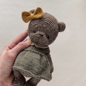 May include: A brown crocheted teddy bear wearing a green dress and a yellow bow. The bear is being held by a hand.