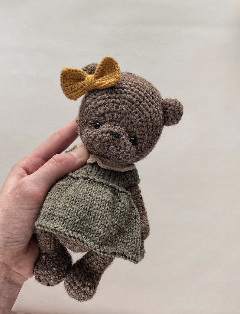 May include: A brown crocheted teddy bear wearing a green dress and a yellow bow. The bear is being held by a hand.