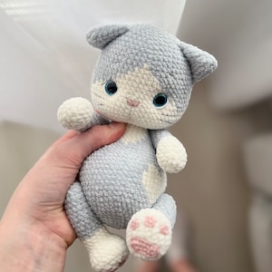 Puede incluir: Un juguete de peluche de gato gris y blanco tejido a crochet con almohadillas de patas rosas. El gato tiene ojos azules y una cara blanca.