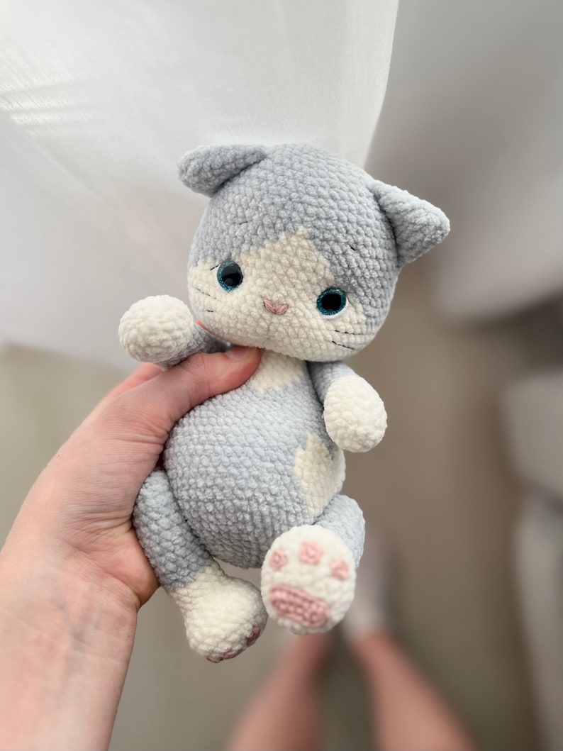 Puede incluir: Un juguete de peluche de gato gris y blanco tejido a crochet con almohadillas de patas rosas. El gato tiene ojos azules y una cara blanca.