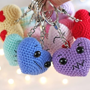 May include: A set of crocheted heart-shaped keychains in various colors, including yellow, green, blue, purple, pink, and red. Each heart has a different facial expression, with embroidered eyes and mouths.