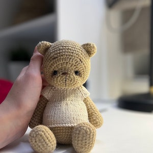 May include: A crocheted teddy bear wearing a white knitted shirt. The bear is sitting on a white surface.