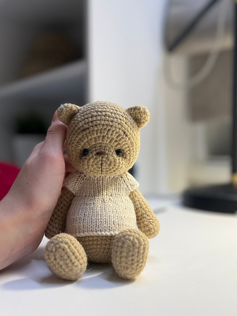 May include: A crocheted teddy bear wearing a white knitted shirt. The bear is sitting on a white surface.