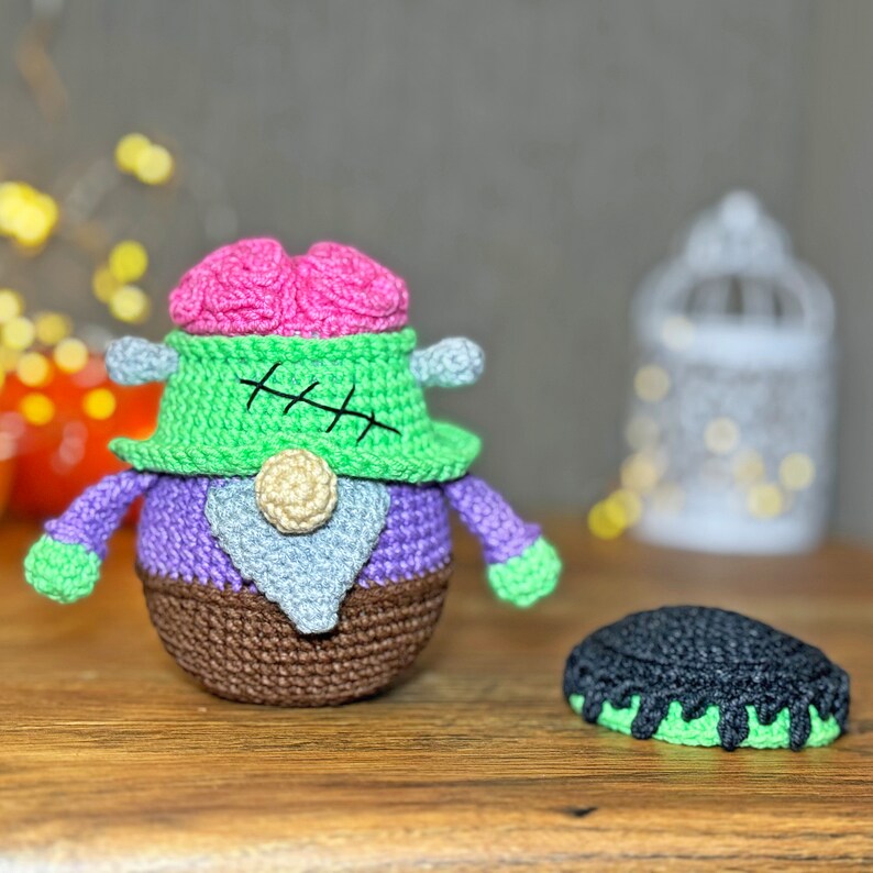 May include: A crocheted plush toy shaped like a Frankenstein monster with a pink brain, green skin, purple body, and brown pants. The toy is sitting on a wooden table next to a black and green crocheted hat.