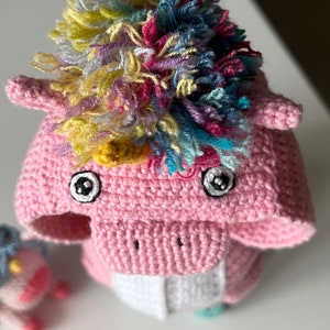 May include: A pink crocheted unicorn toy with a rainbow mane and large black eyes. The unicorn is standing on a white surface.