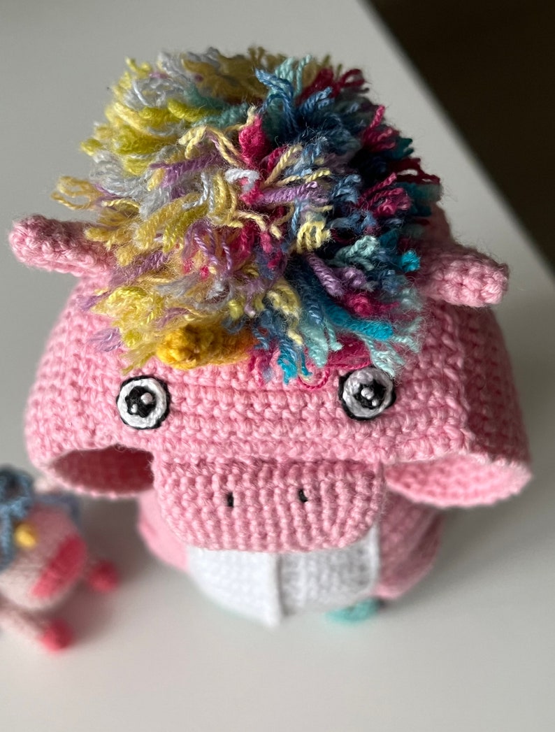 May include: A pink crocheted unicorn toy with a rainbow mane and large black eyes. The unicorn is standing on a white surface.