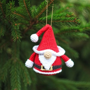 May include: A crocheted Santa Claus ornament with a red hat, white beard, and black belt. The ornament is hanging from a tree branch.