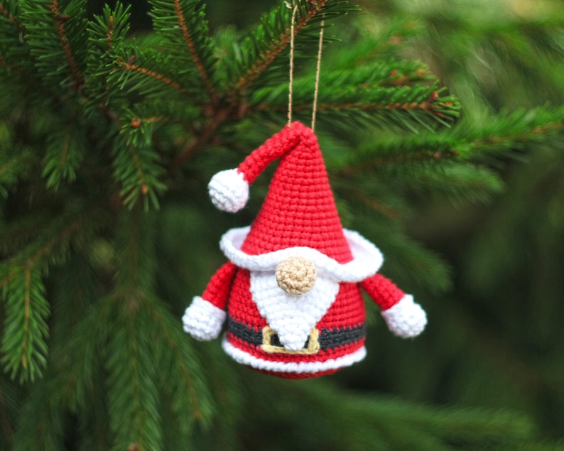 May include: A crocheted Santa Claus ornament with a red hat, white beard, and black belt. The ornament is hanging from a tree branch.