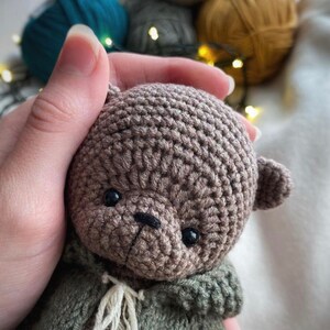 May include: A crocheted brown teddy bear with black eyes and a black nose. The bear is wearing a green crocheted jacket with a white crocheted scarf.