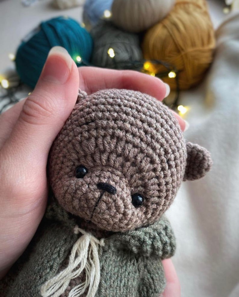 May include: A crocheted brown teddy bear with black eyes and a black nose. The bear is wearing a green crocheted jacket with a white crocheted scarf.