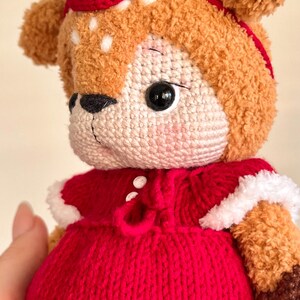May include: A handmade, plush deer doll with a red knitted dress and matching hat. The deer has brown fur, white spots, and large black eyes. The dress is trimmed with white faux fur.