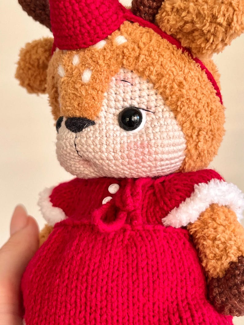 May include: A handmade, plush deer doll with a red knitted dress and matching hat. The deer has brown fur, white spots, and large black eyes. The dress is trimmed with white faux fur.