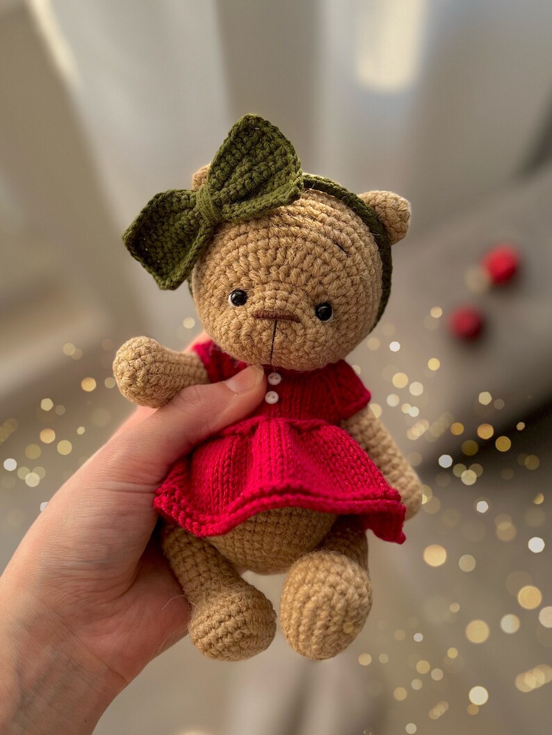 May include: A crocheted teddy bear wearing a red dress and a green bow headband. The bear is being held by a hand.