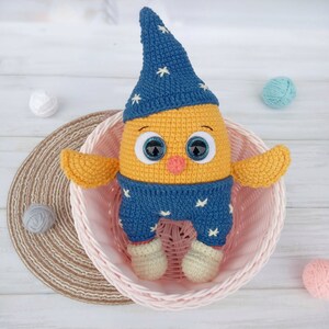 May include: A crocheted yellow chick wearing a blue and white star patterned hat and pajamas. The chick has large, sparkly eyes and a small orange nose. The chick is sitting in a pink wicker basket.