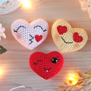 May include: Three crocheted hearts with different facial expressions. The first heart is pink with closed eyes and a red heart on its mouth. The second heart is yellow with red hearts for eyes and a smile. The third heart is red with a wink, a black eye, and a red tongue.