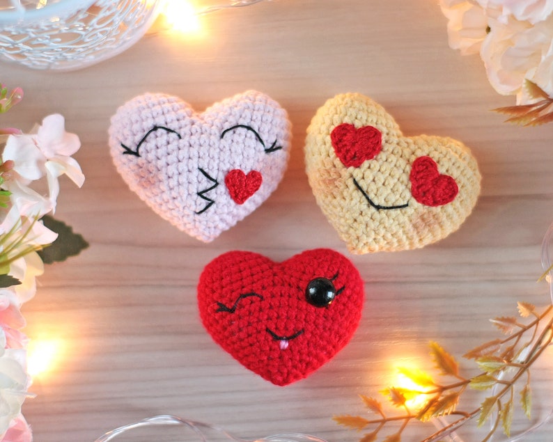 May include: Three crocheted hearts with different facial expressions. The first heart is pink with closed eyes and a red heart on its mouth. The second heart is yellow with red hearts for eyes and a smile. The third heart is red with a wink, a black eye, and a red tongue.