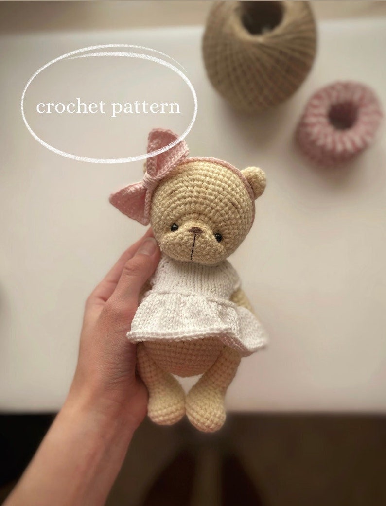 May include: A crocheted teddy bear wearing a white dress and a pink bow. The bear is light brown and has black eyes.