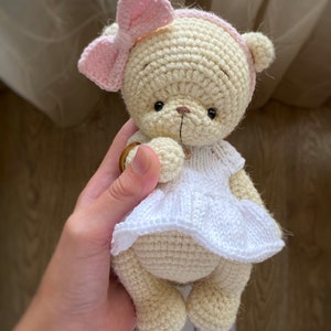 May include: A crocheted teddy bear wearing a white dress and a pink bow. The bear is being held by a hand.