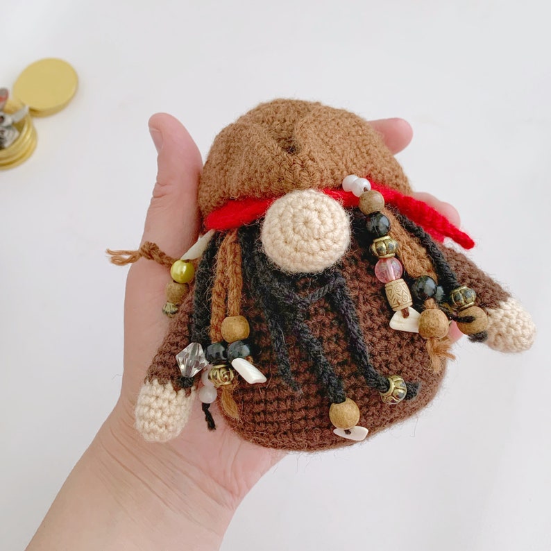 May include: A crocheted gnome with brown hair, a red bandana, and a beige beard. The gnome is decorated with brown, black, white, and gold beads.