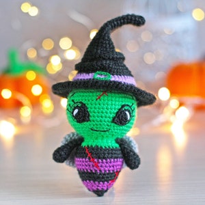 May include: A handmade crochet Halloween decoration featuring a green alien-like creature wearing a black witch hat with purple trim. The creature has black and purple striped body, small wings, and stitched details. Pumpkins and fairy lights are in the background.