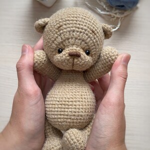May include: A crocheted brown teddy bear with black eyes and a brown nose. The bear is being held by two hands.