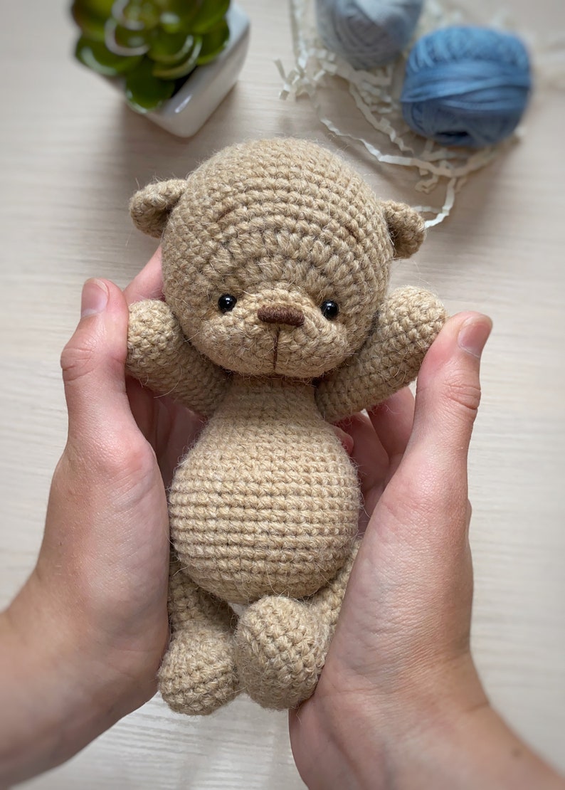 May include: A crocheted brown teddy bear with black eyes and a brown nose. The bear is being held by two hands.