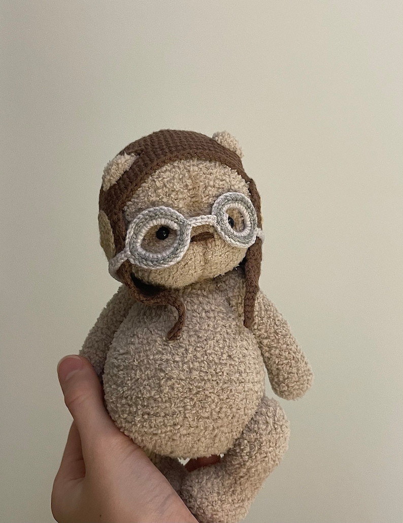 May include: A crocheted plush toy bear wearing a brown crocheted hat with ear flaps and round, gray-rimmed glasses.