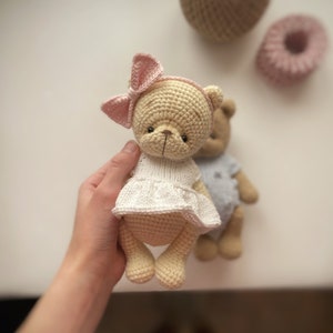 May include: Two crocheted teddy bears, one wearing a white dress and a pink bow, the other wearing a blue dress. The bears are made with beige yarn and have black eyes and noses.