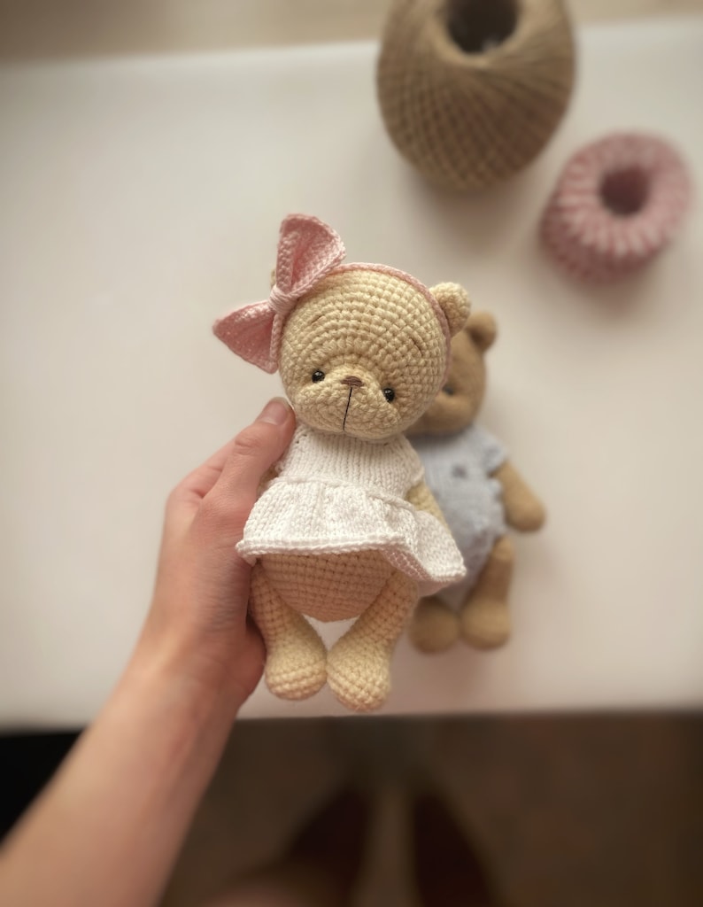 May include: Two crocheted teddy bears, one wearing a white dress and a pink bow, the other wearing a blue dress. The bears are made with beige yarn and have black eyes and noses.