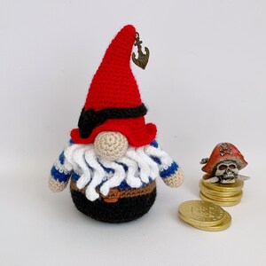 May include: A crocheted gnome wearing a red hat with a black eye patch and a blue and white striped shirt. The gnome is holding a small anchor.